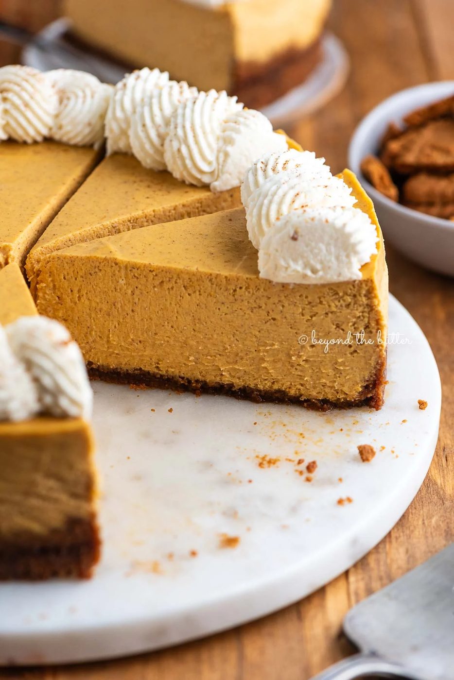 Sliced pumpkin cheesecake topped with piped whipped cream on a white marbled cake plate on a dark wood background.