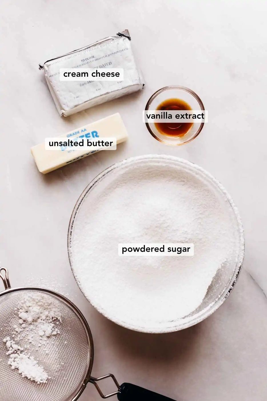 Ingredients in various size bowls labeled as cream cheese, vanilla extract, unsalted butter, and powdered sugar on a white marbled background.