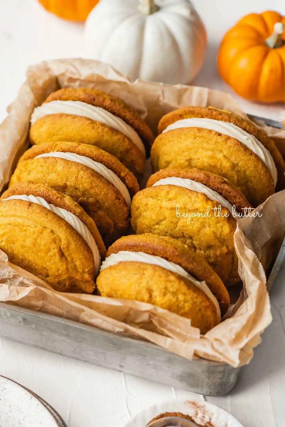 Six pumpkin whoopie pies placed in a brown parchment paper lined metal baking tin with mini orange and white pumpkins placed around it on a white background.