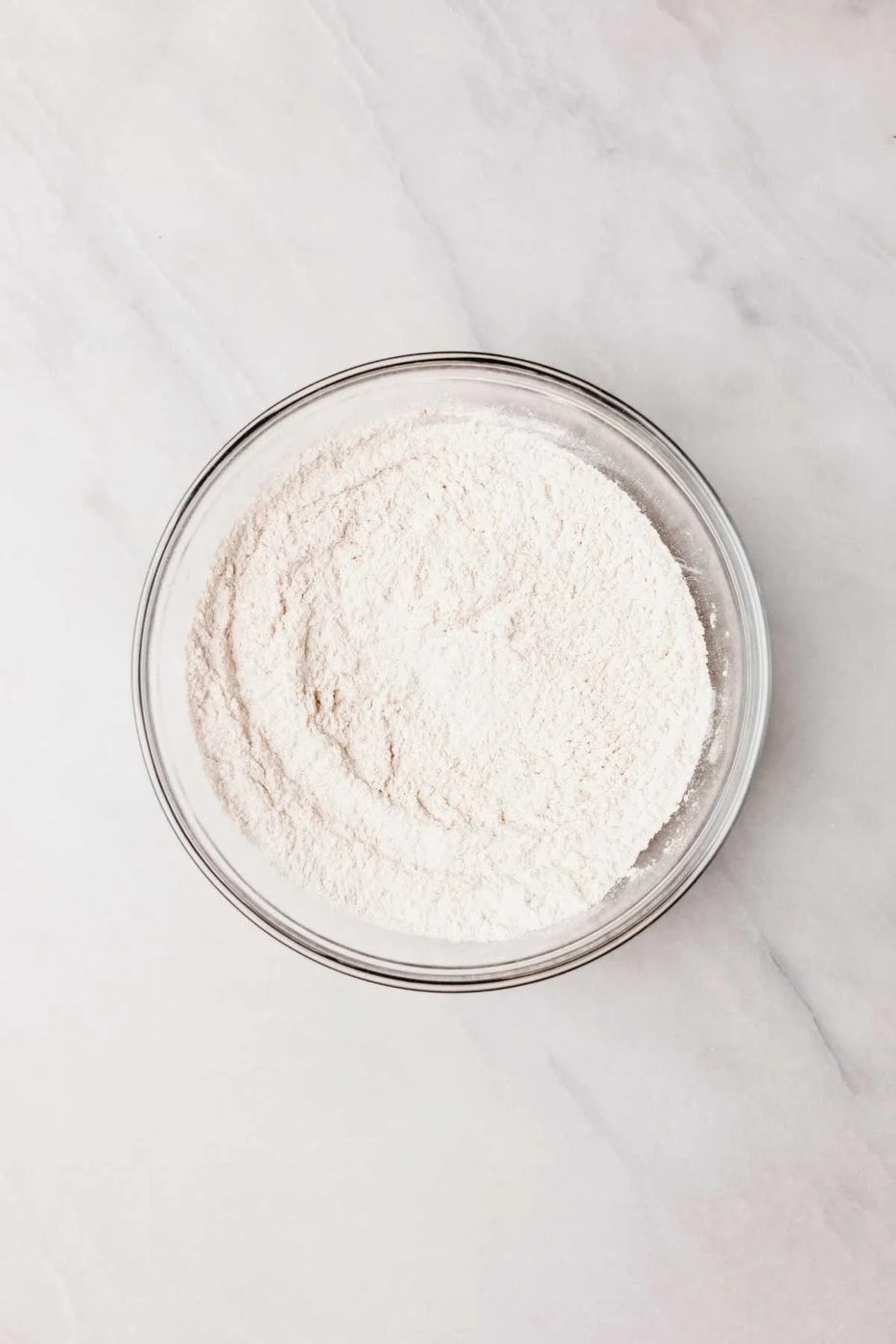Whisked all purpose flour, baking soda, baking powder, cinnamon, and salt in glass mixing bowl on light grey marbled background.
