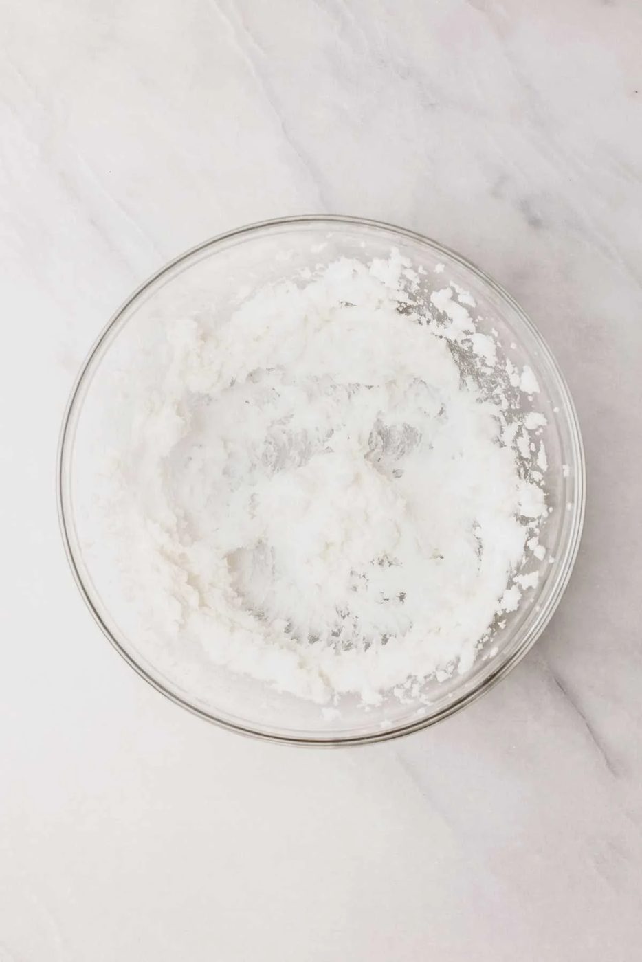 Vegetable shortening and granulated sugar beaten together in a large glass mixing bowl on a light grey marbled background.
