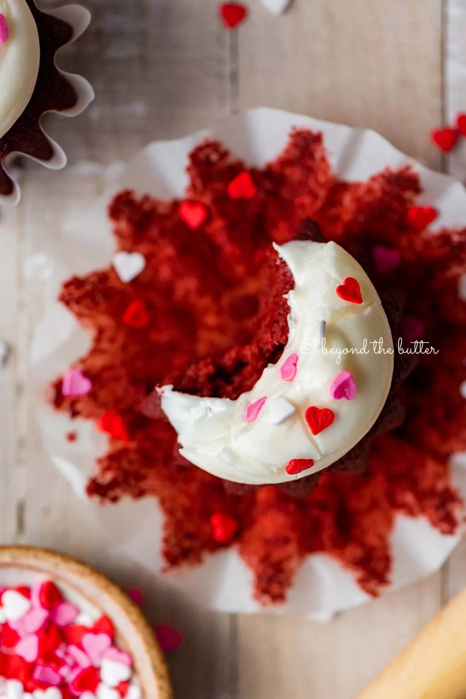 House-made red velvet cupcakes with one opened and half eaten with a small bowl of Valentine's Day sprinkles on light wood background | All images © Sovuh