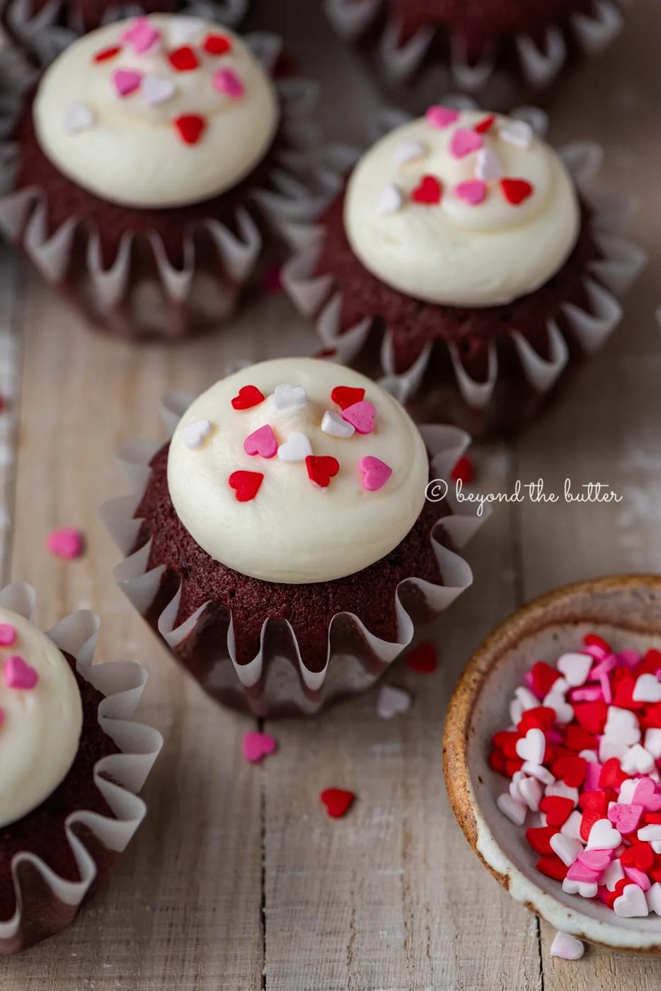 House-made red velvet cupcakes with cream cheese frosting topped with heart sprinkles on a light wood background | All images © Sovuh