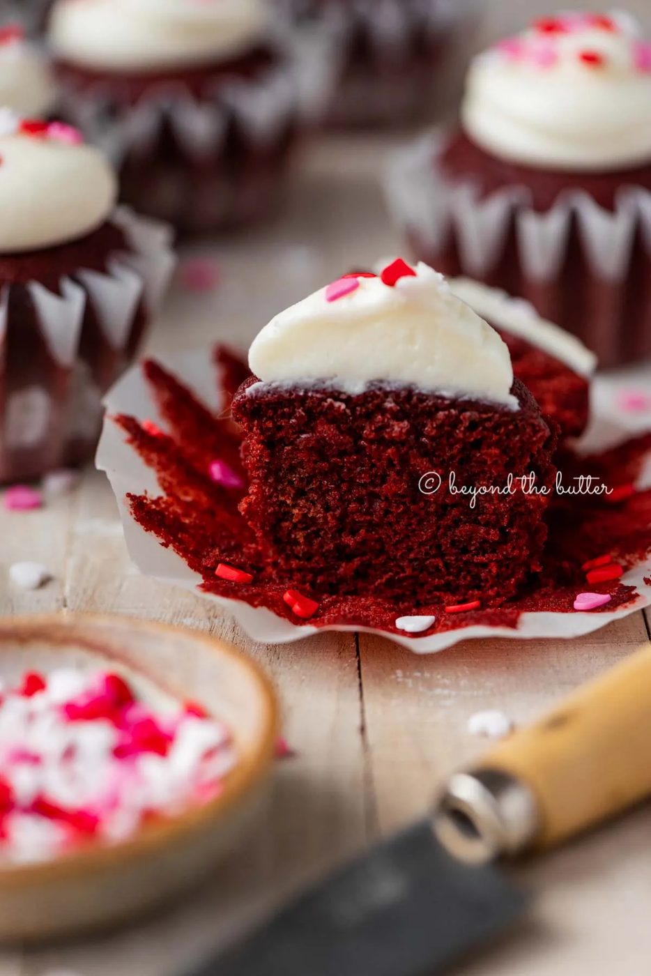 Sliced red velvet cupcake with cream cheese frosting topped with heart sprinkles on a light wood background | All images © Sovuh