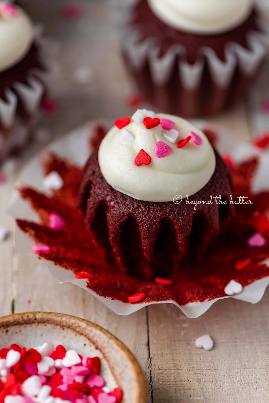 Opened red velvet cupcake with cream cheese frosting topped with heart sprinkles on a light wood background | All images © Sovuh