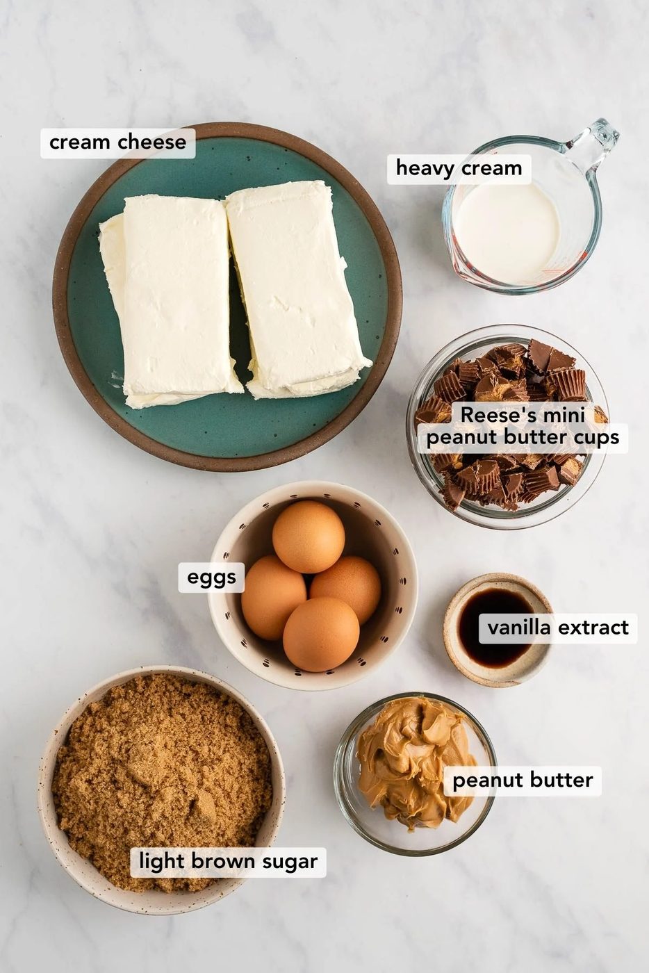 Overhead image of ingredients used to make Reese's peanut butter cup cheesecake on white and grey marble background.