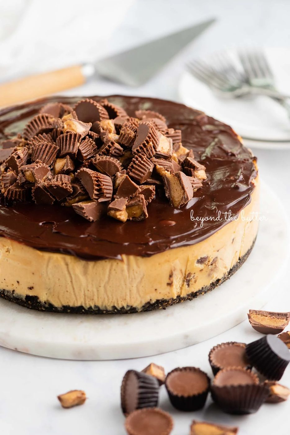 Peanut butter cheesecake topped with chocolate ganache and mini Reese's peanut butter cups on marble plate on white background.