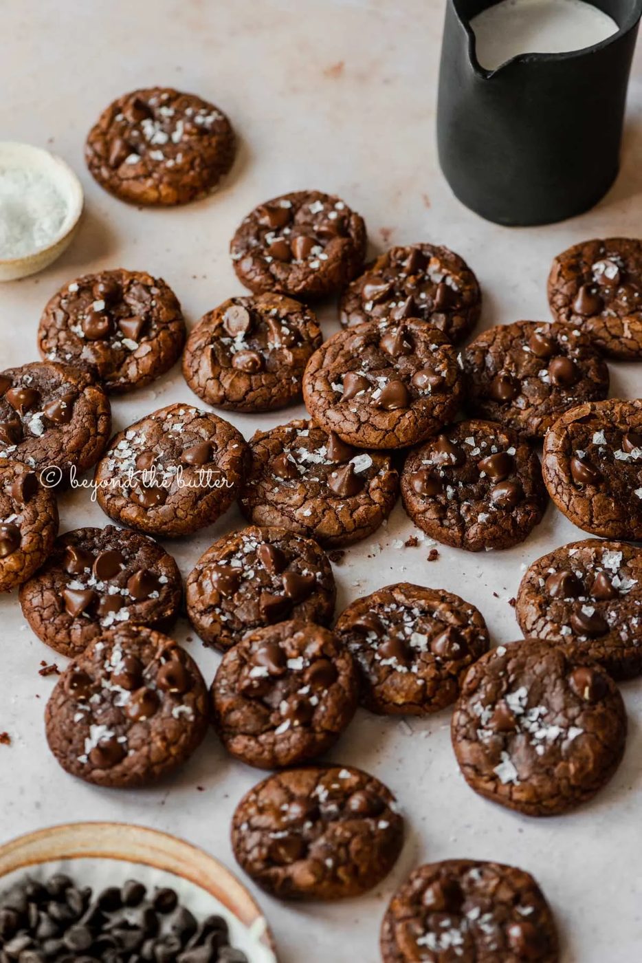 Salted chocolate chip brownie cookies randomly placed on an abstract light brown background with small black pitcher of milk and bowl of sea salt flakes to the side | All Images © Sovuh