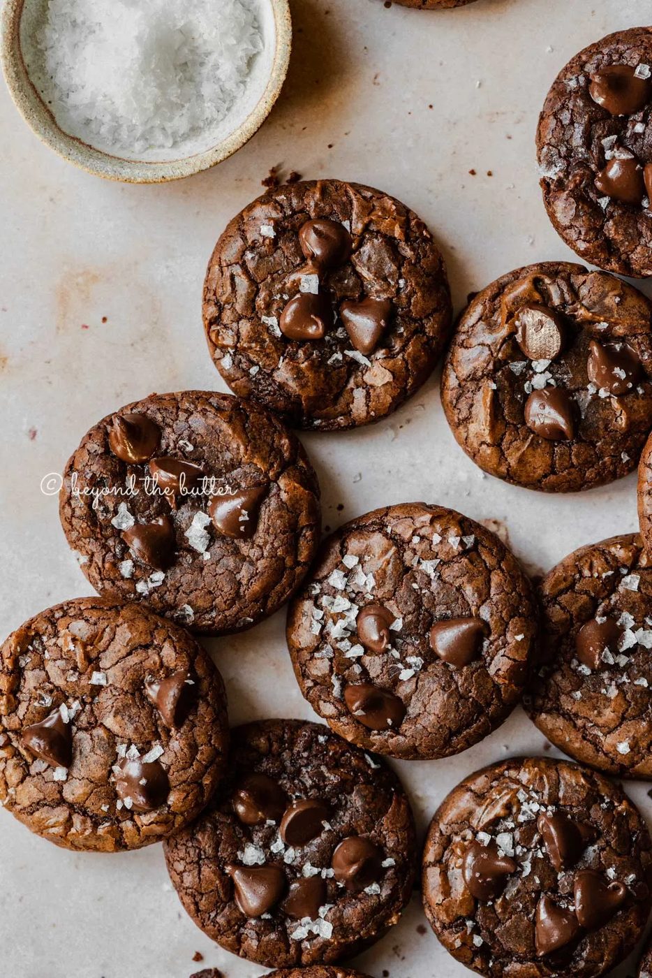 Salted chocolate chip brownie cookies randomly placed on an abstract light brown background with small bowl of sea salt flakes to the side | All Images © Sovuh