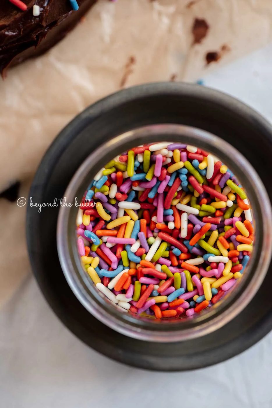 Small bowl of rainbow sprinkles on a dark plate with chocolate cake next to it | All Images © Sovuh