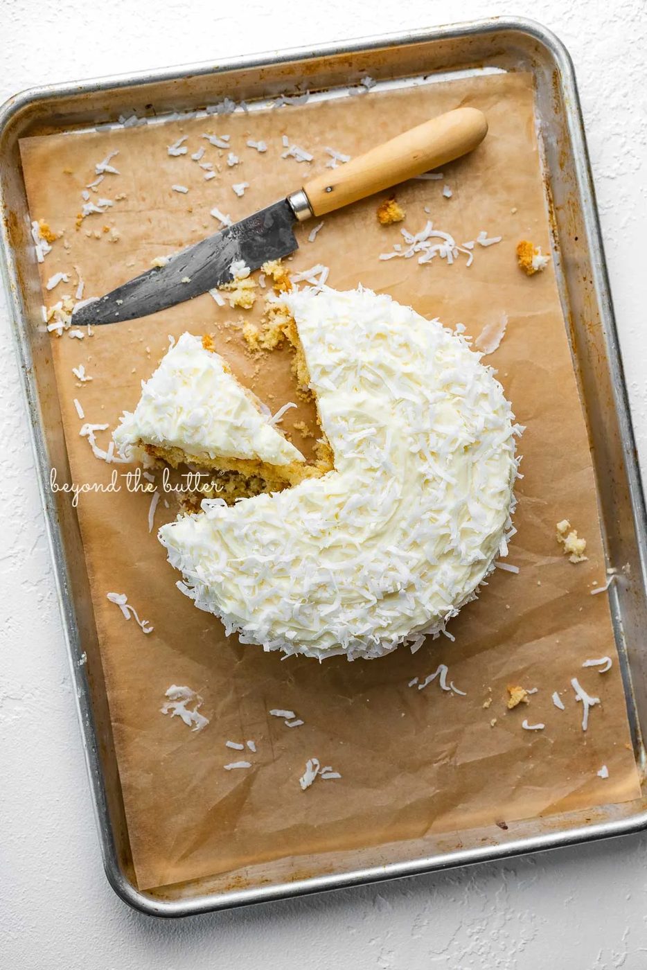 Single layer coconut cake frosted with coconut buttercream with slice cut out on natural parchment paper-lined baking sheet | © Sovuh