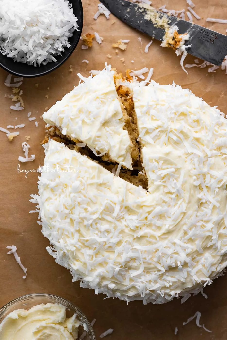 Single layer coconut cake frosted with coconut buttercream with slice cut out on natural parchment paper background | © Sovuh