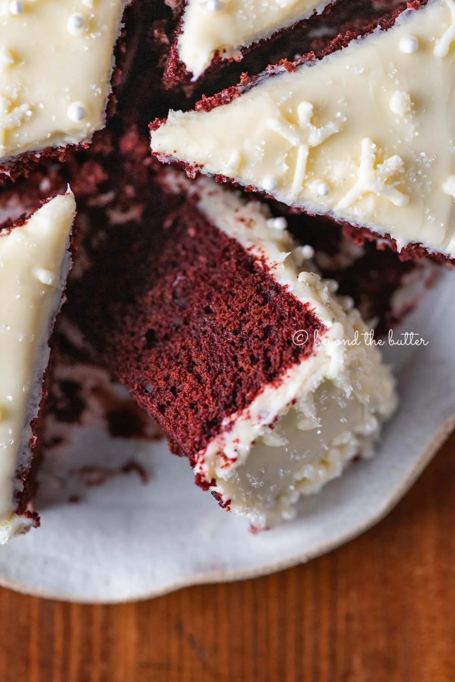 Sliced red velvet cake with cream cheese frosting on a scalloped edge dessert plate with red plaid background | All Images © Sovuh