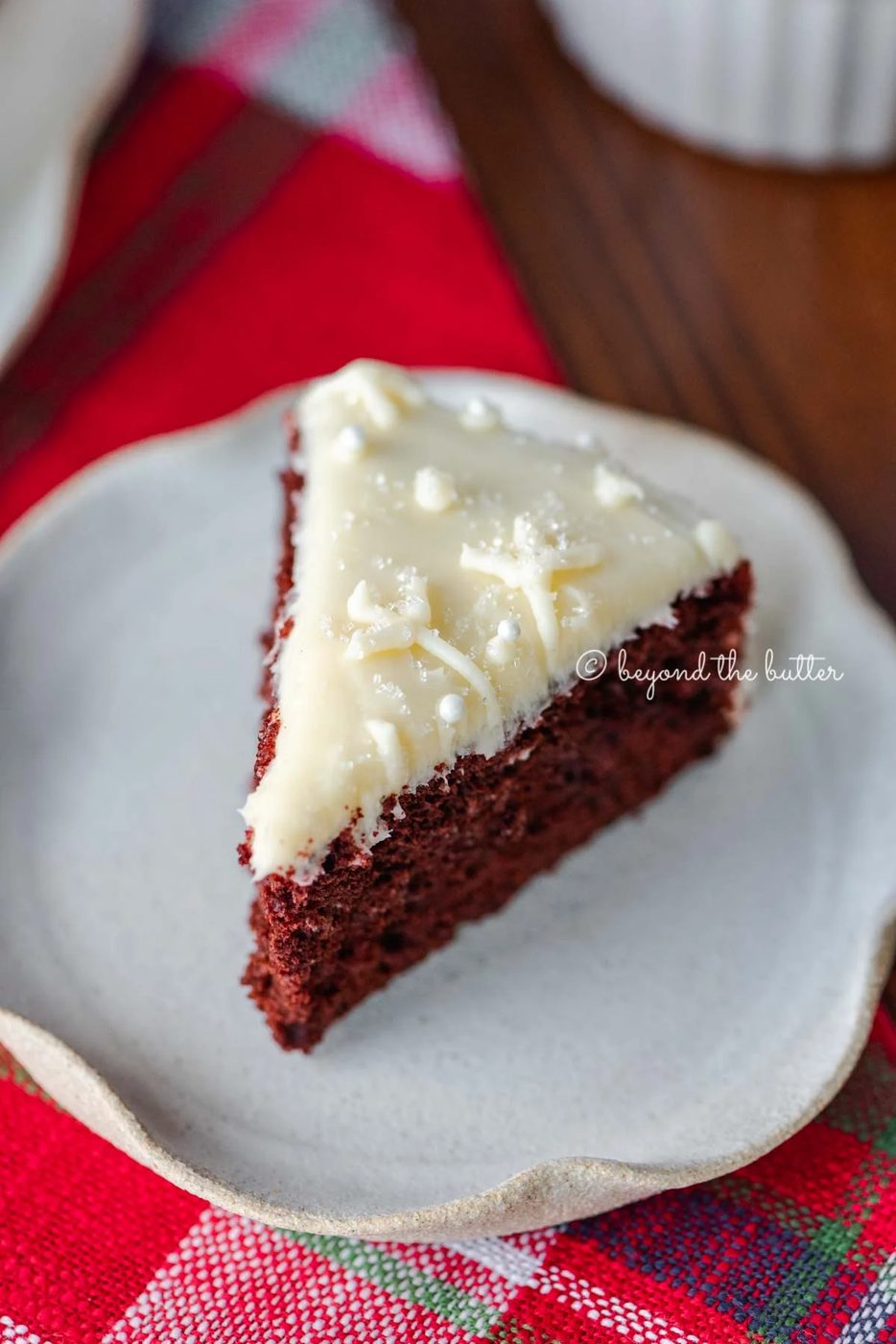 Slice of red velvet cake with cream cheese frosting on a scalloped edge dessert plate with red plaid background | All Images © Sovuh