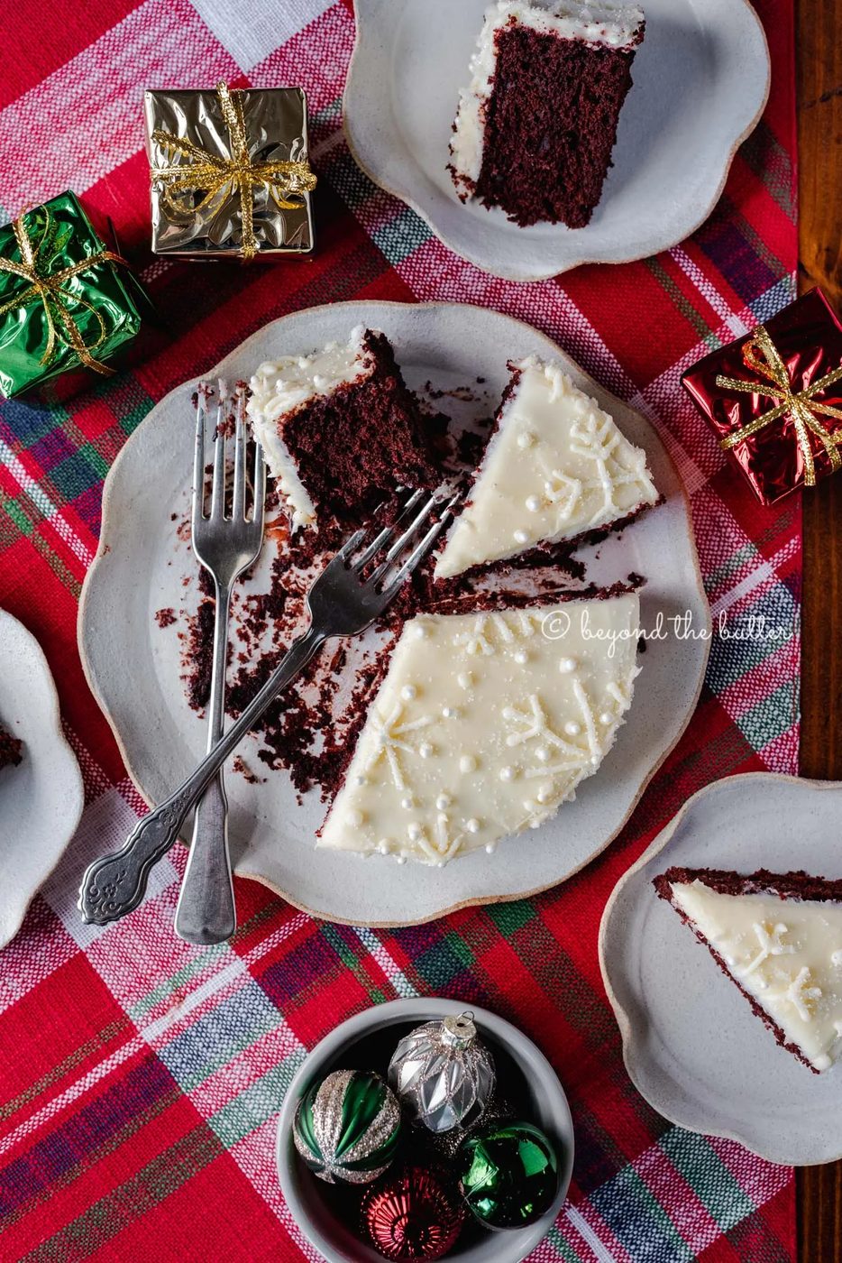 Half eaten and sliced red velvet cake with cream cheese frosting on a scalloped edge dessert plate with red plaid background | All Images © Sovuh