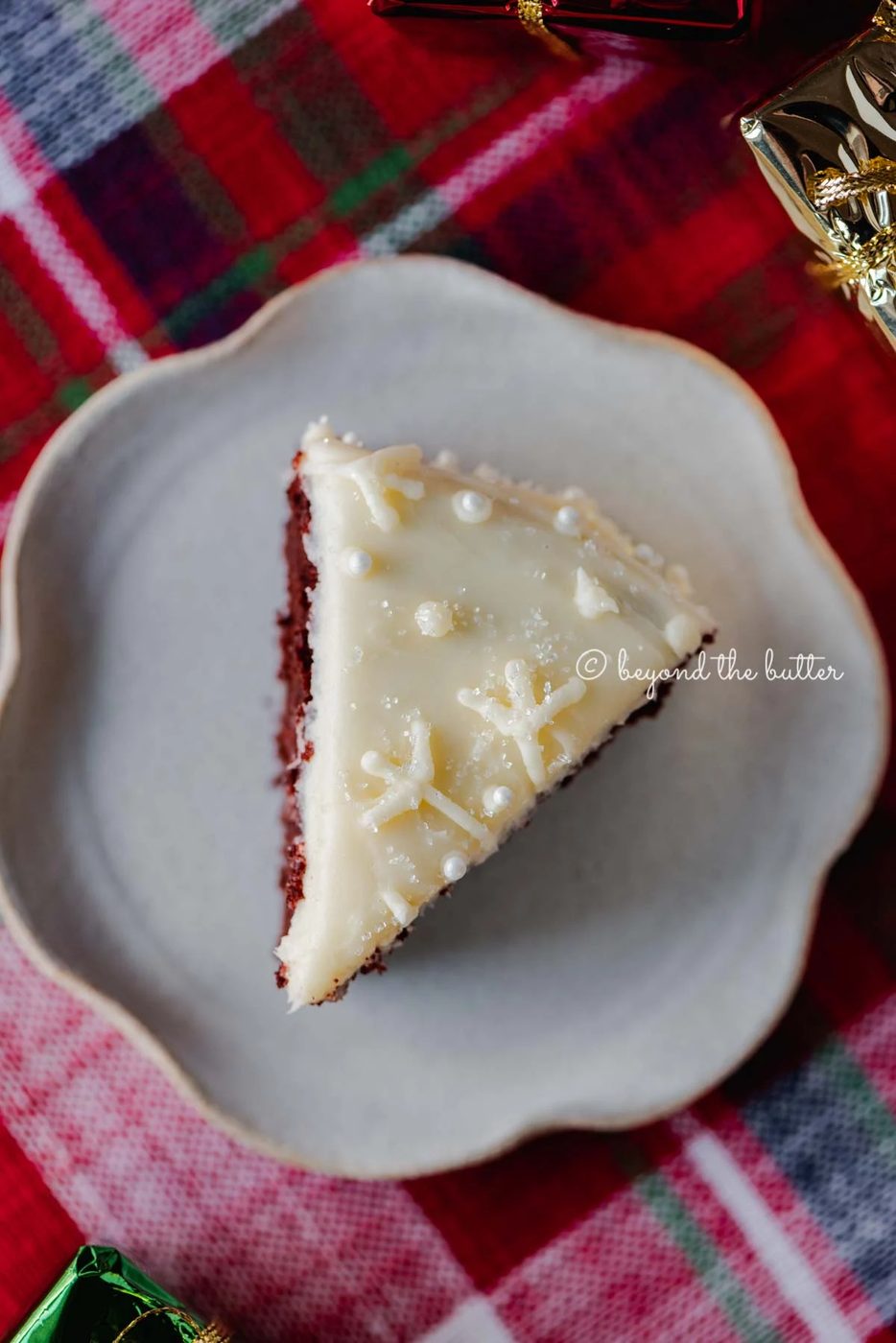 Slice of red velvet cake with cream cheese frosting on a scalloped edge dessert plate with red plaid background | All Images © Sovuh