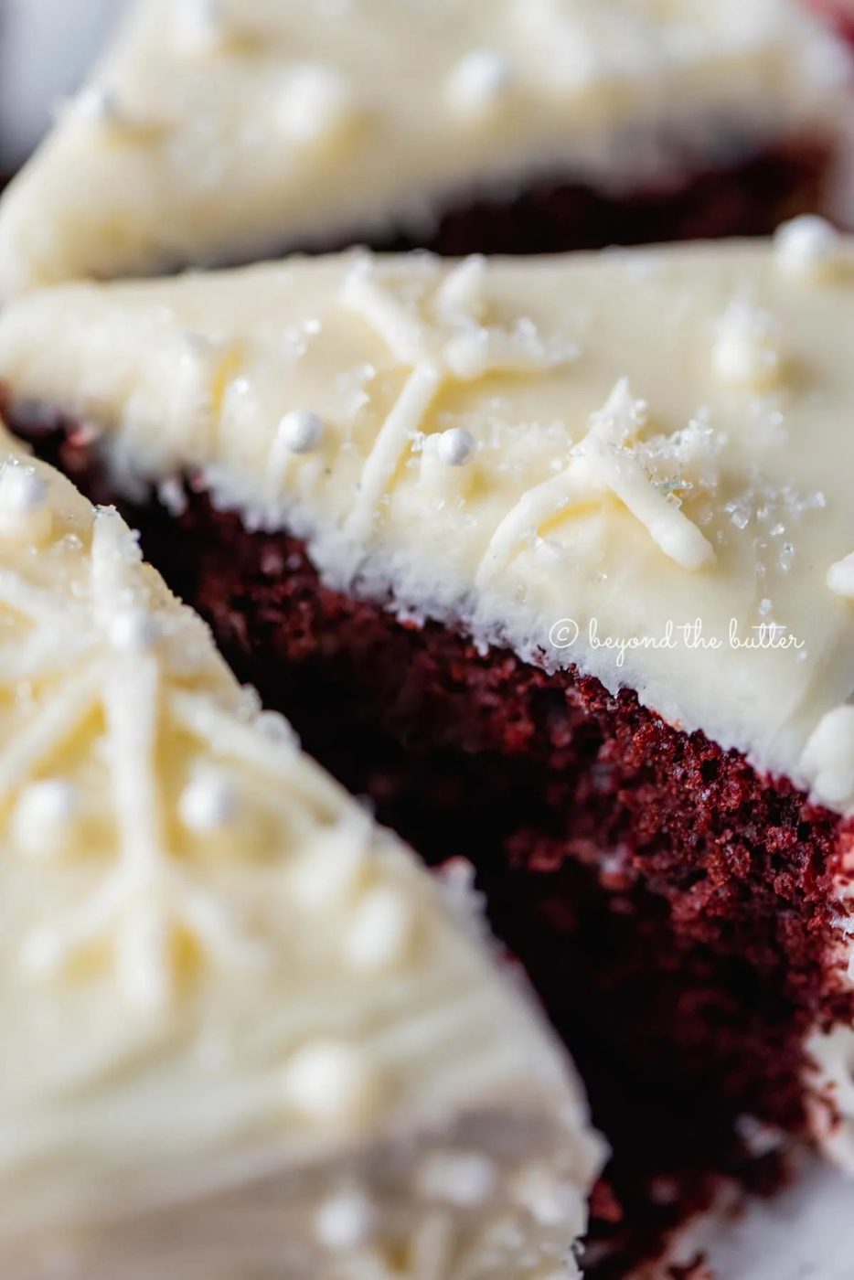 Closeup of sliced red velvet cake with cream cheese frosting on a scalloped edge dessert plate with red plaid background | All Images © Sovuh