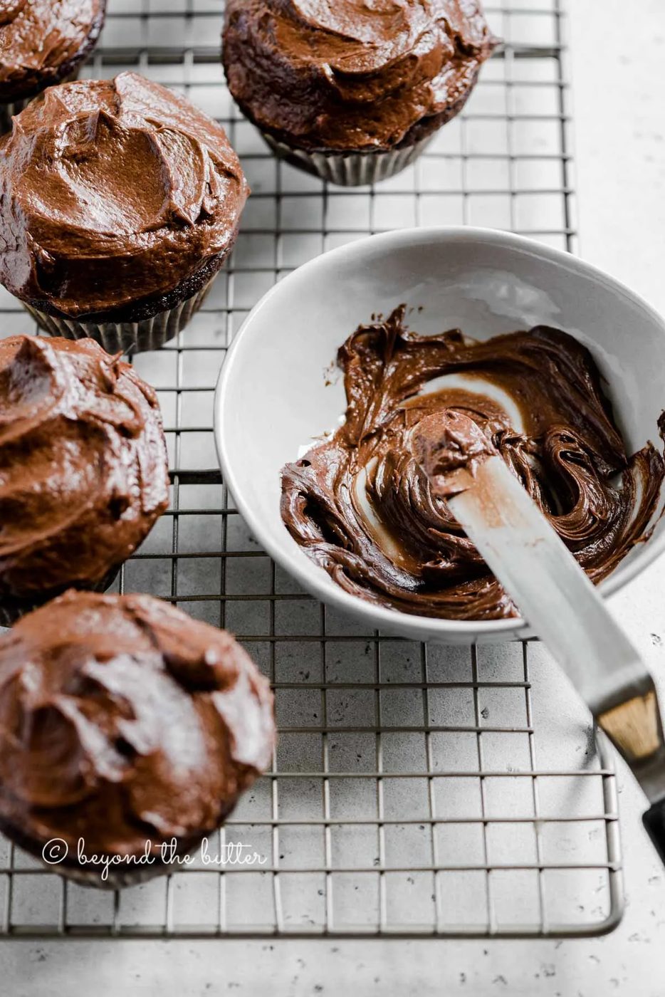 Angled image of chocolate cupcakes on cooling rack with small bowl of chocolate frosting and offset spatula | All Images © Sovuh