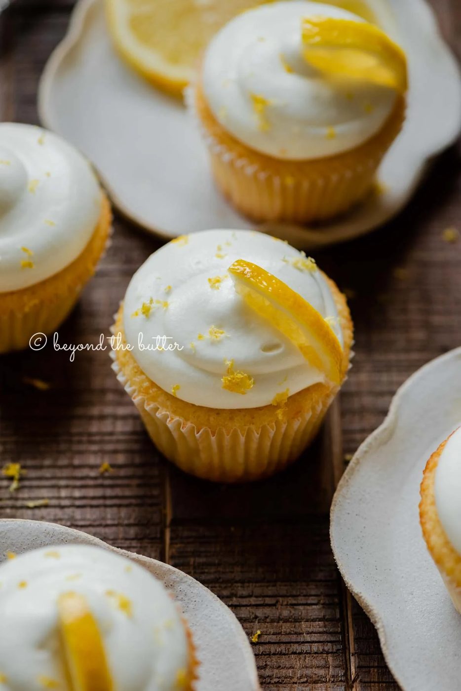 Randomly placed lemon cupcakes with lemon cream cheese frosting, garnished with lemon triangles with a small bowl of lemon zest on a dark wood background | All Images © Sovuh