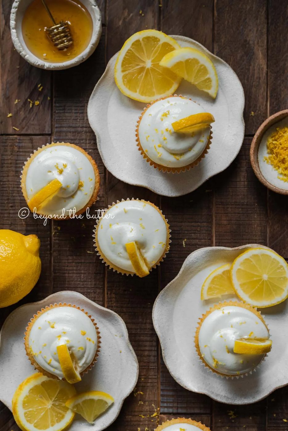 Randomly placed lemon cupcakes with lemon cream cheese frosting, garnished with lemon triangles on a dark wood background | All Images © Sovuh