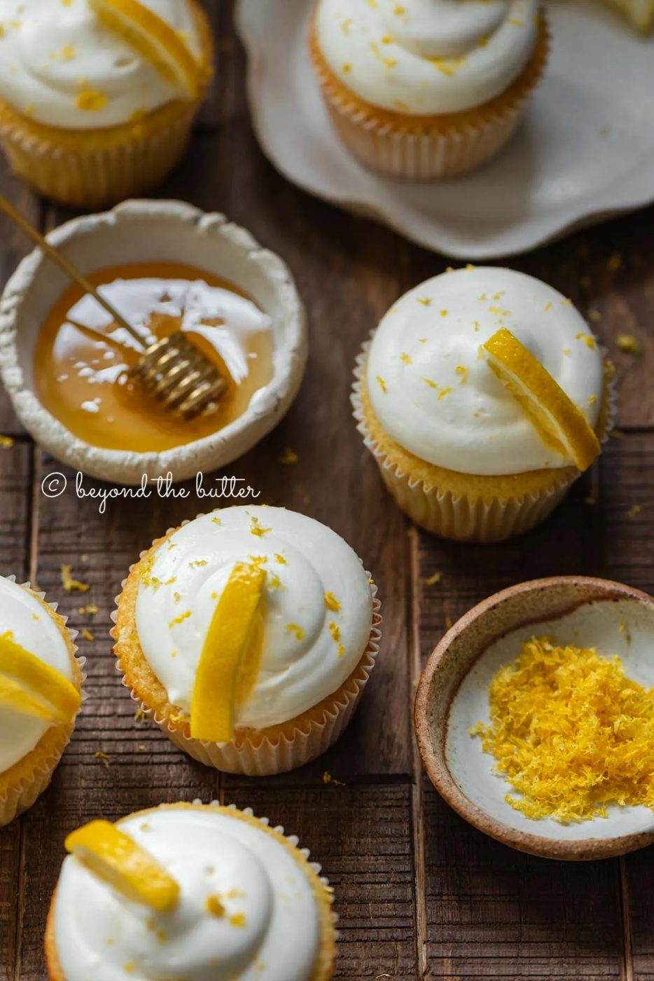 Randomly placed lemon cupcakes with a small bowl of honey and small bowl of lemon zest on a dark wood background | All Images © Sovuh