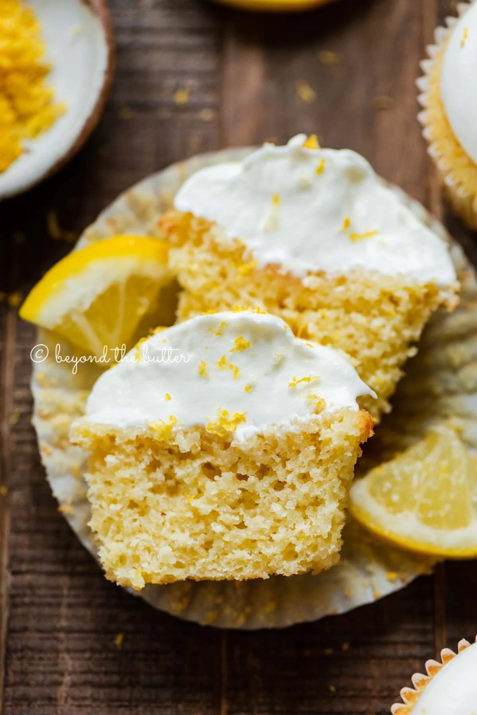 Sliced open lemon cupcake with whipped lemon cream cheese frosting on cupake liner and 2 wedges of lemons on a dark wood background | All Images © Sovuh