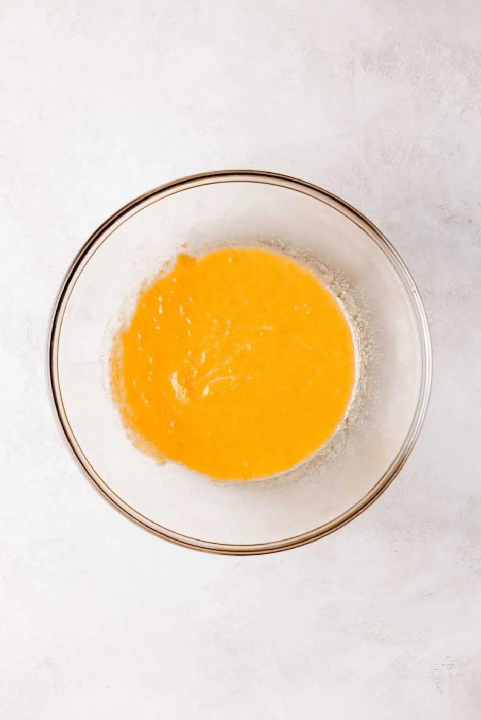 Whisked milk, pumpkin puree, melted butter, and egg in a medium size glass mixing bowl on a white and grey marbled background.