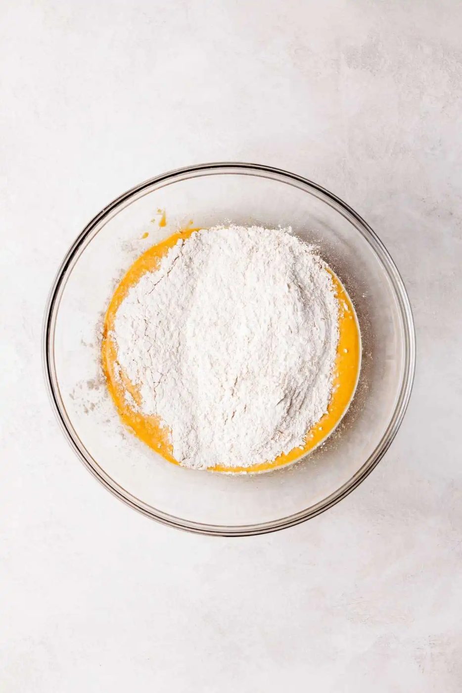 Flour mixture added to pumpkin cinnamon roll wet ingredients in a medium size glass mixing bowl on a white and grey marbled background.