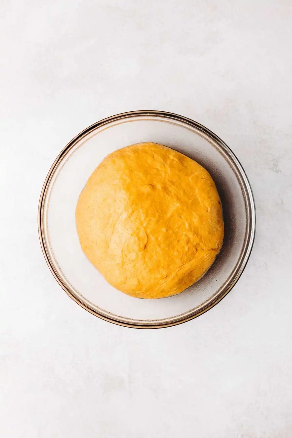 Pumpkin cinnamon roll dough doubled in size in a medium size glass mixing bowl on a white and grey marbled background.