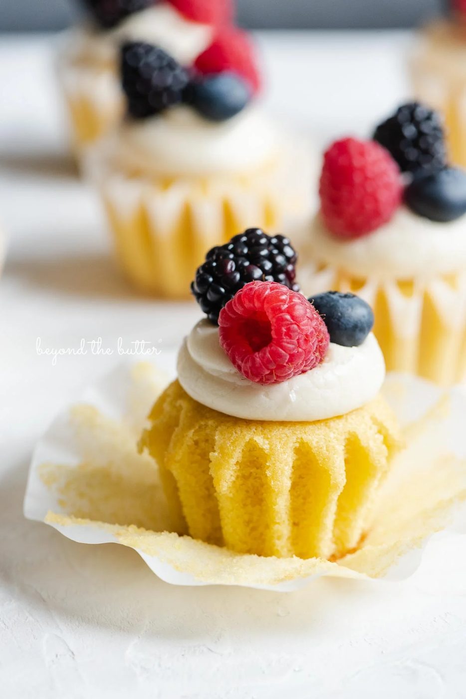 Angled image of small batch vanilla cupakes topped with vanilla buttercream frosting and berries with front cupcake unwrapped.