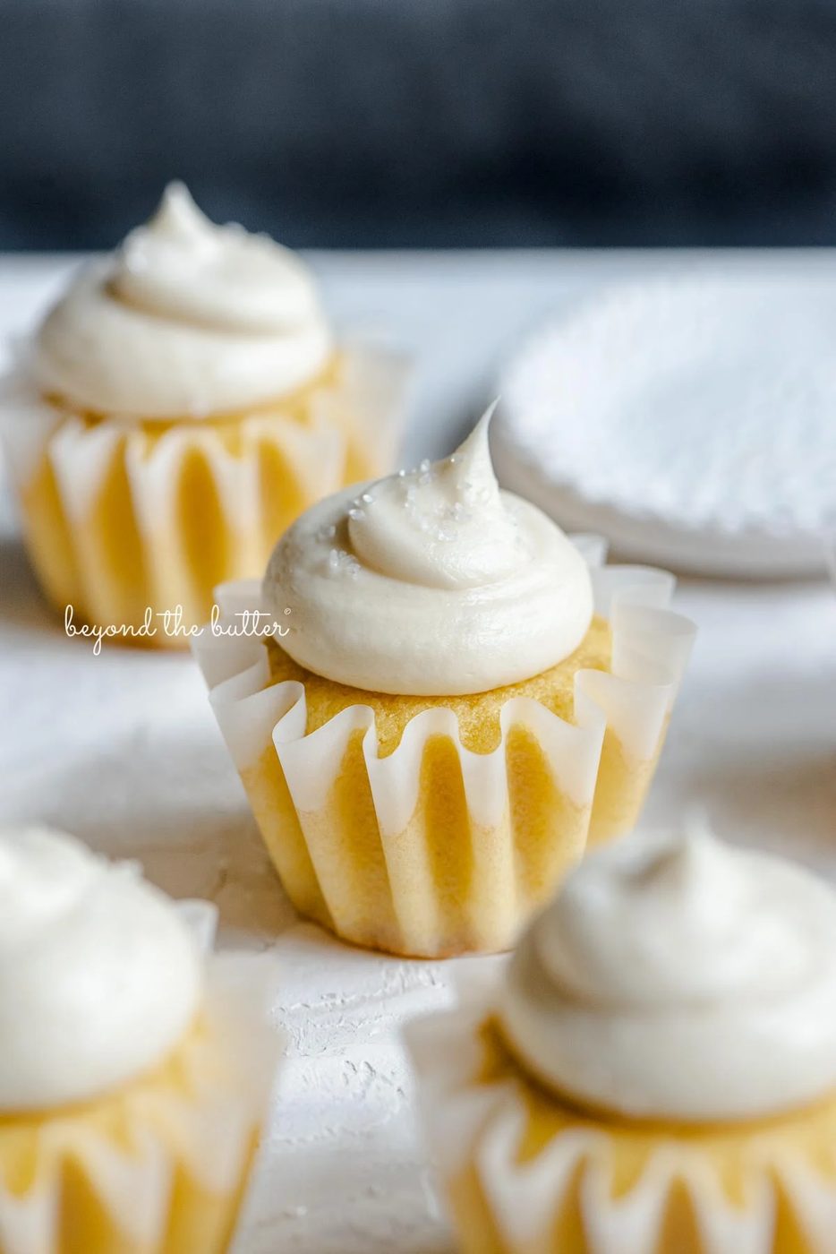 Angled image of frosted small batch vanilla cupcakes sprinkled with sparkling sugar randomly placed on white and dark blue background next to a small shallow white bowl of sparkling sugar.