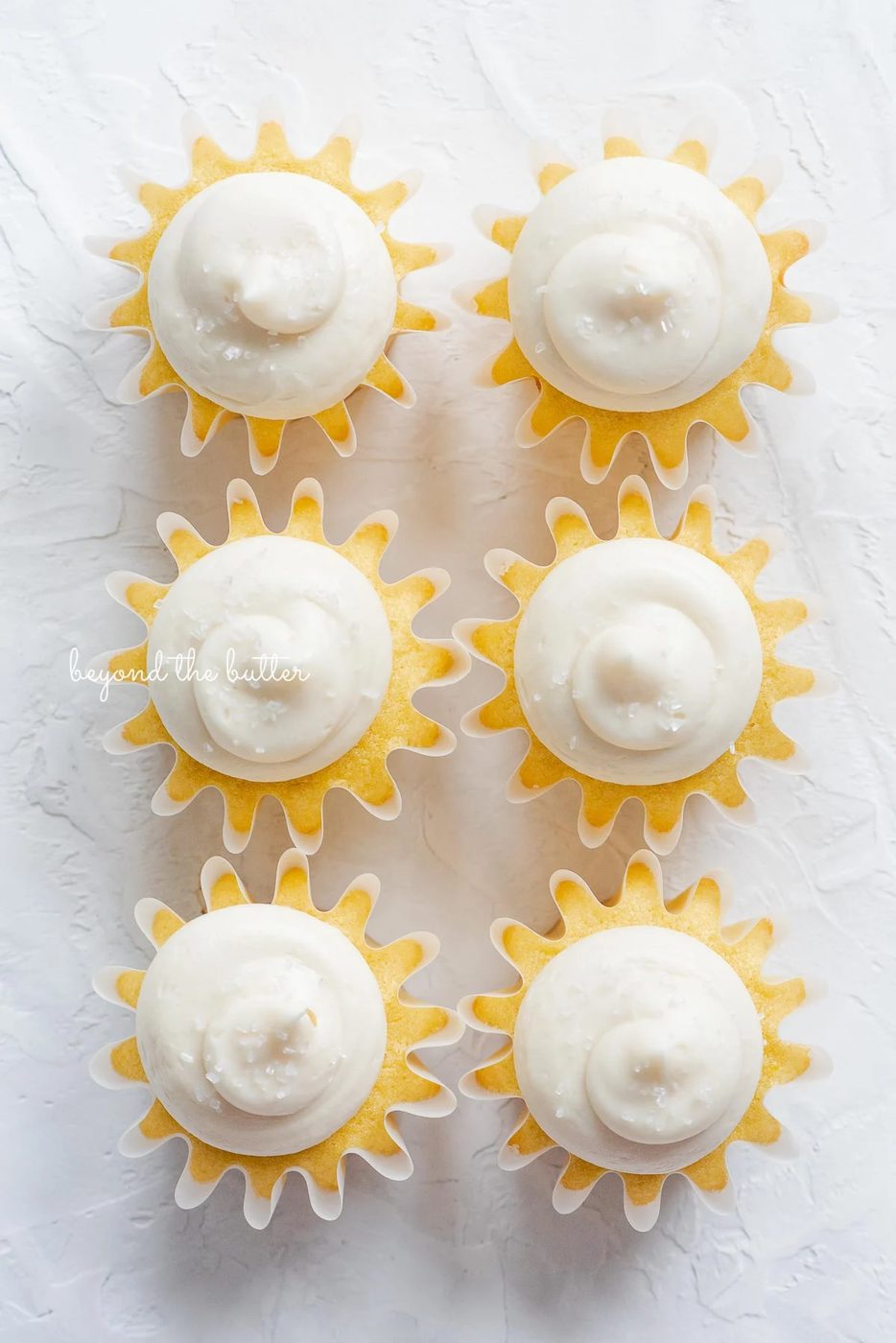 Overhead image of six small batch vanilla cupcakes with vanilla buttercream frosting sprinkled with sparkling sugar in two columns on white background.