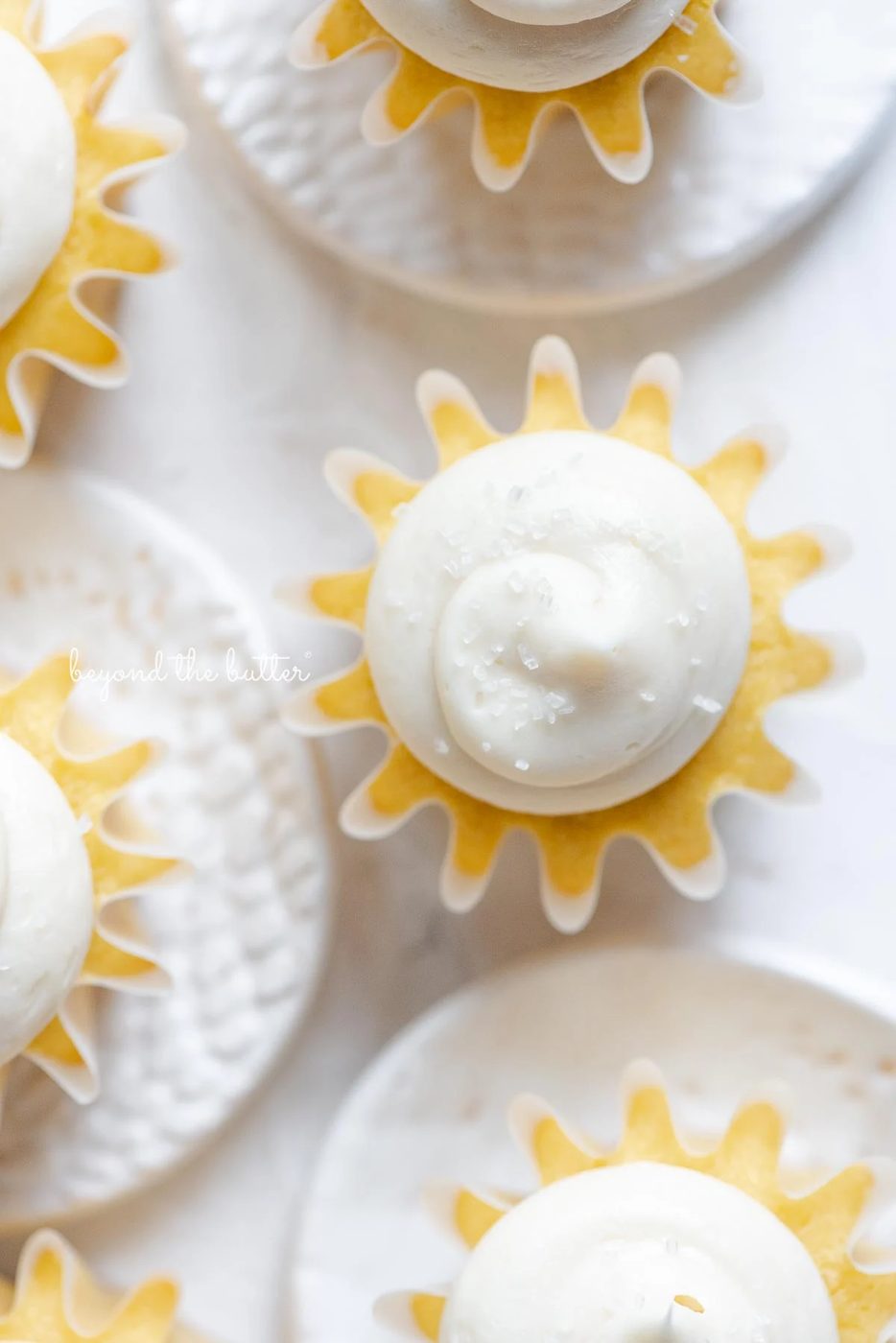 Overhead image of vanilla cupcakes with vanilla buttercream frosting sprinkled with sparkling sugar on white background and small ceramic plates.