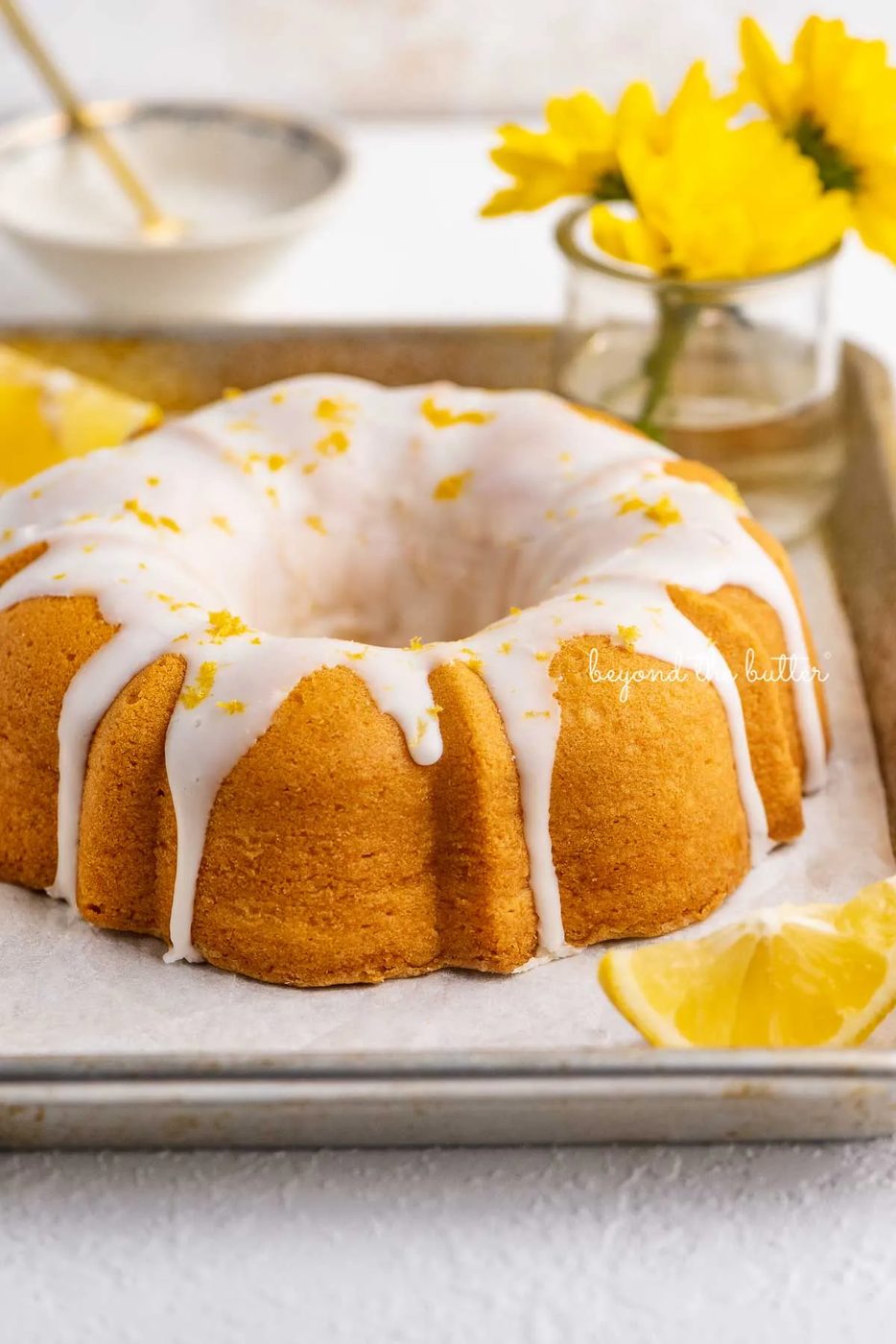 Small lemon cream cheese pound cake topped with a simple lemon glaze on a parchment paper lined baking sheet with sliced lemons and small bouquet of daisies | © Sovuh