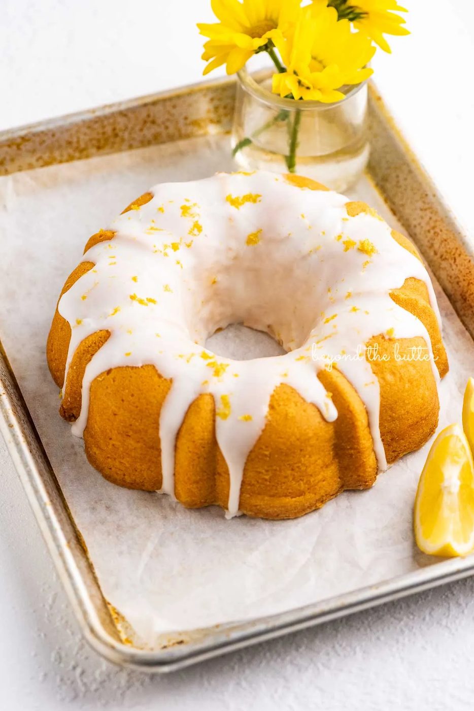 Small lemon cream cheese pound cake topped with a simple lemon glaze on a parchment paper lined baking sheet with sliced lemons and small bouquet of daisies | © Sovuh