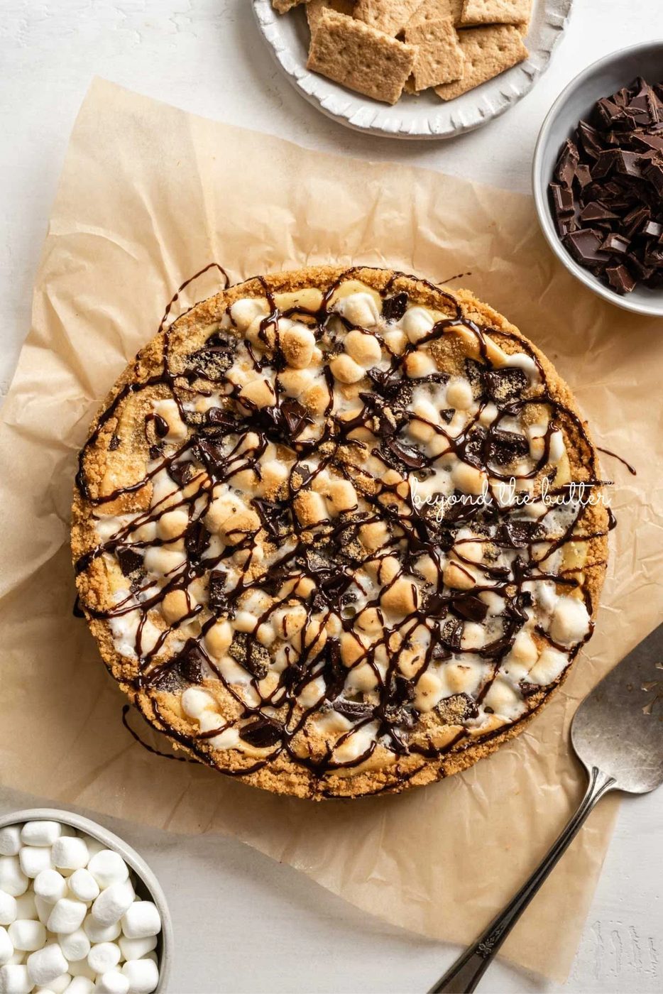 S'mores cheesecake topped with drizzled chocolate syrup placed on a sheet of brown parchment paper with a bowl of mini marshmallows, graham crackers, chocolate, and a cake server placed around it.