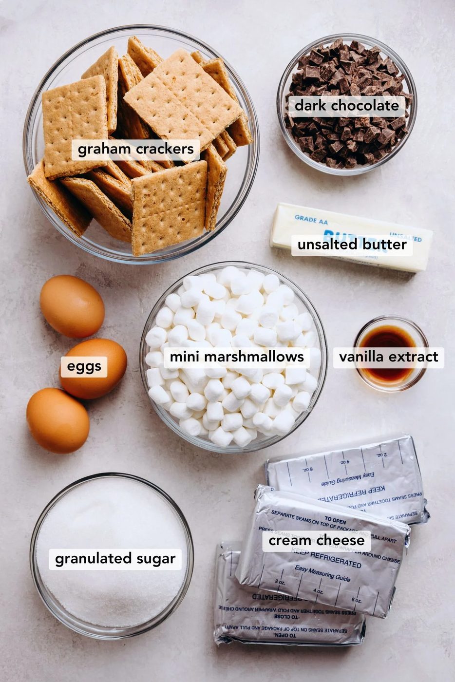 Ingredients in various size bowls labeled as graham crackers, dark chocolate, eggs, mini marshmallows, unsalted butter, vanilla extract, granulated sugar, and cream cheese on a white marbled background.