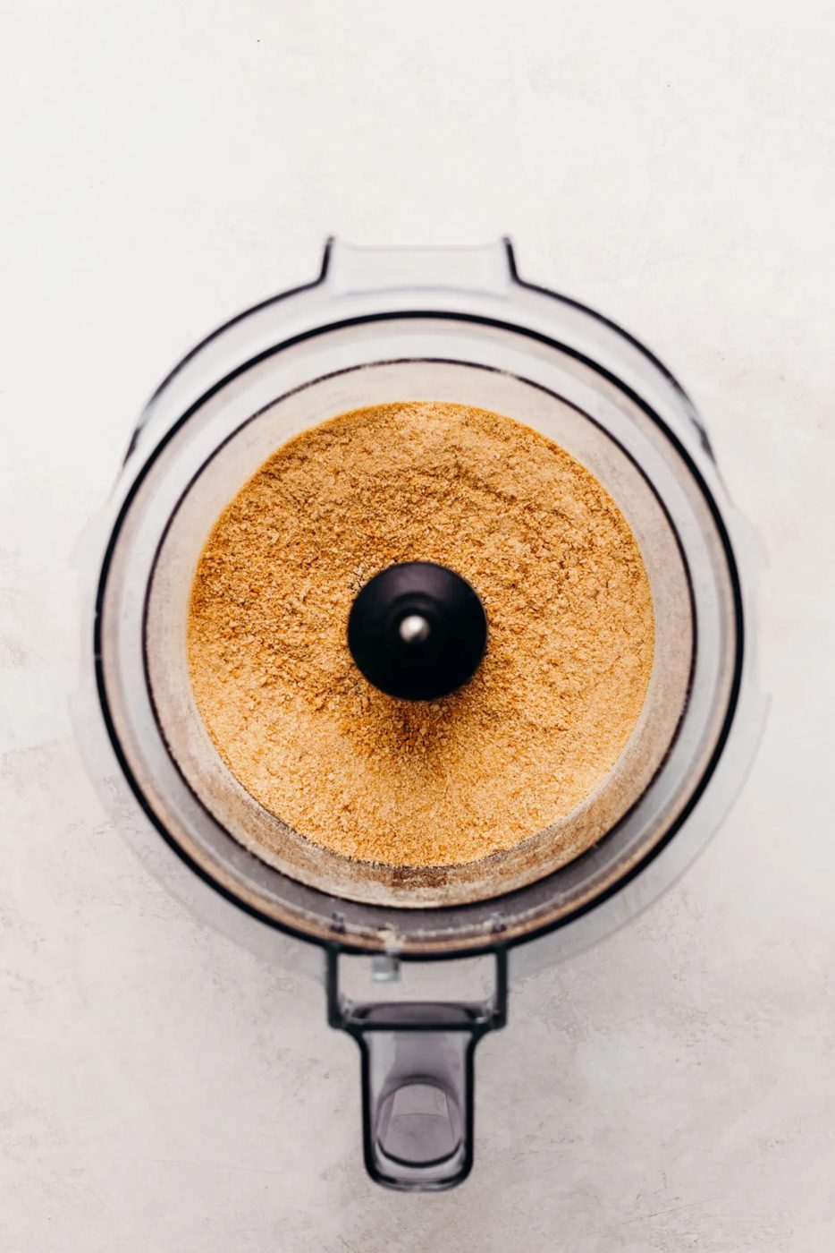 Graham cracker crumbs in a food processor on a textured white background.