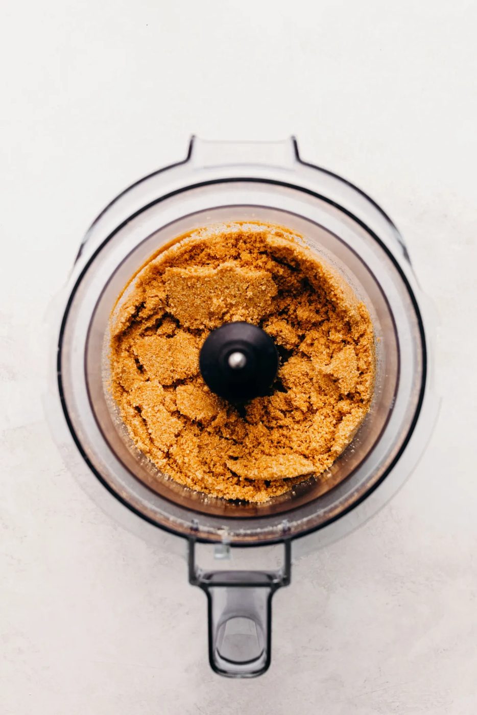 Graham cracker crumbs mixed together with melted butter and sugar in a food processor on a textured white background.