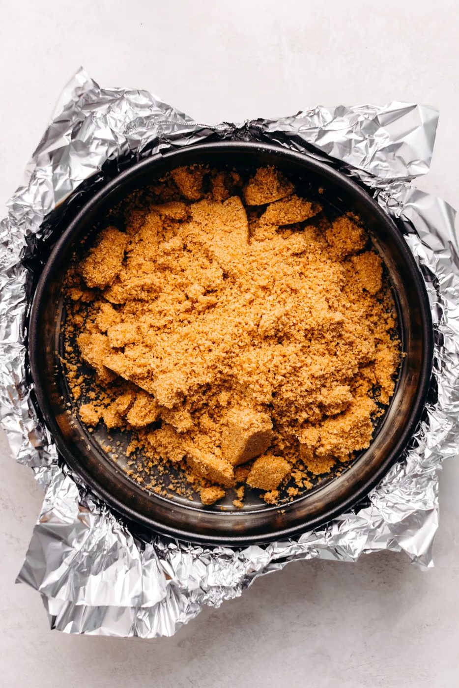 Graham cracker crust mixture placed on a aluminum foil wrapped springform pan on a textured white background.