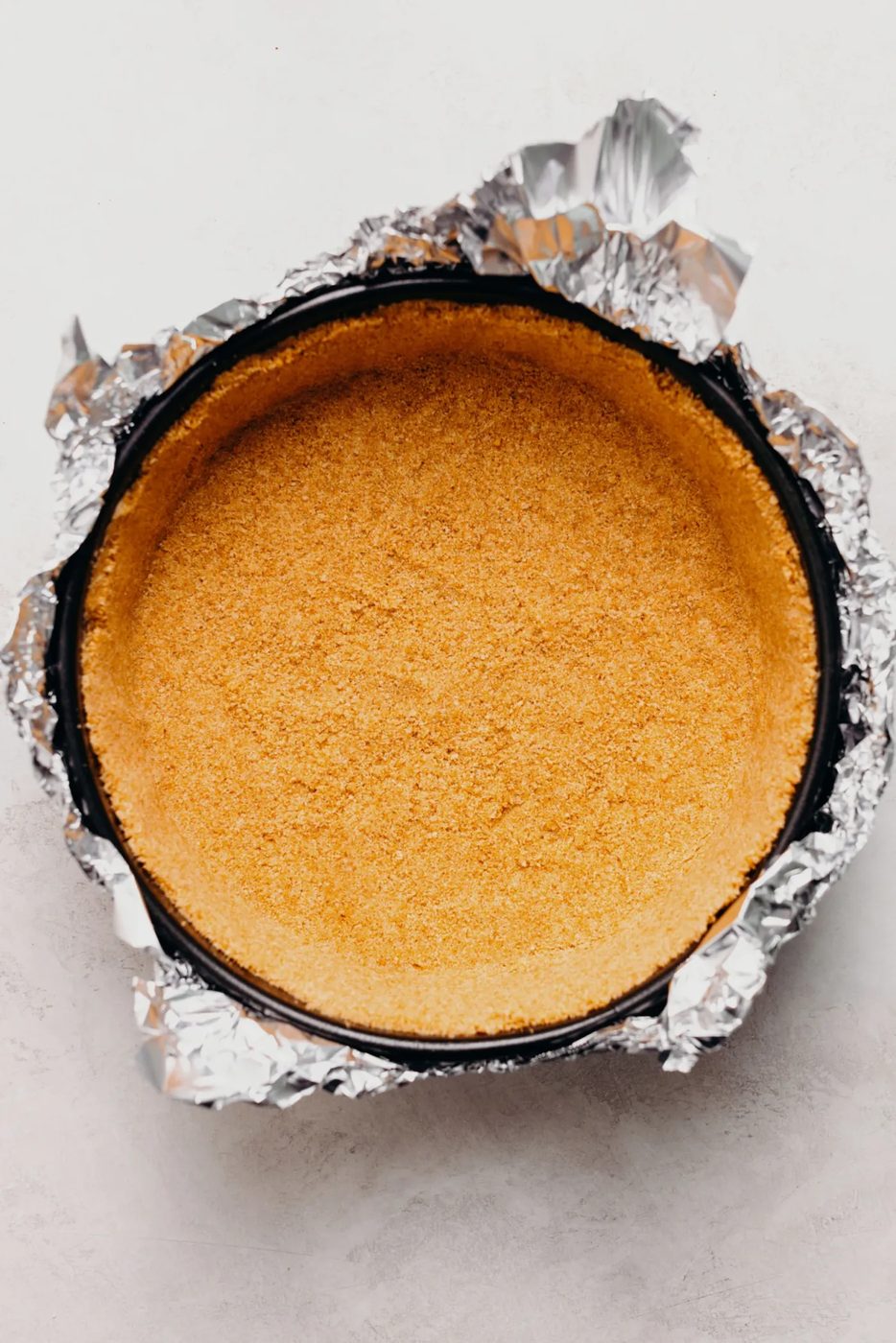 Baked graham cracker crust in a aluminum foil wrapped springform pan on a textured white background.