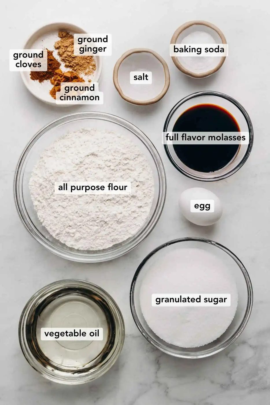 Ingredients in various size bowls labeled as ground cloves, ground ginger, ground cinnamon, salt, baking soda, all purpose flour, full flavor molasses, egg, vegetable oil, and granulated sugar on a white marbled background.