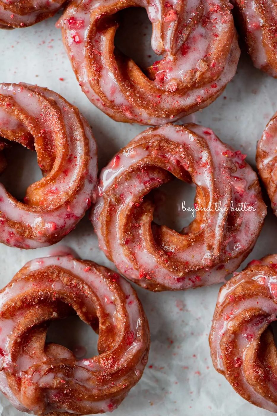 Strawberry glazed french crullers sprinkled with strawberry sugar on a parchment paper lined baking sheet | © Sovuh