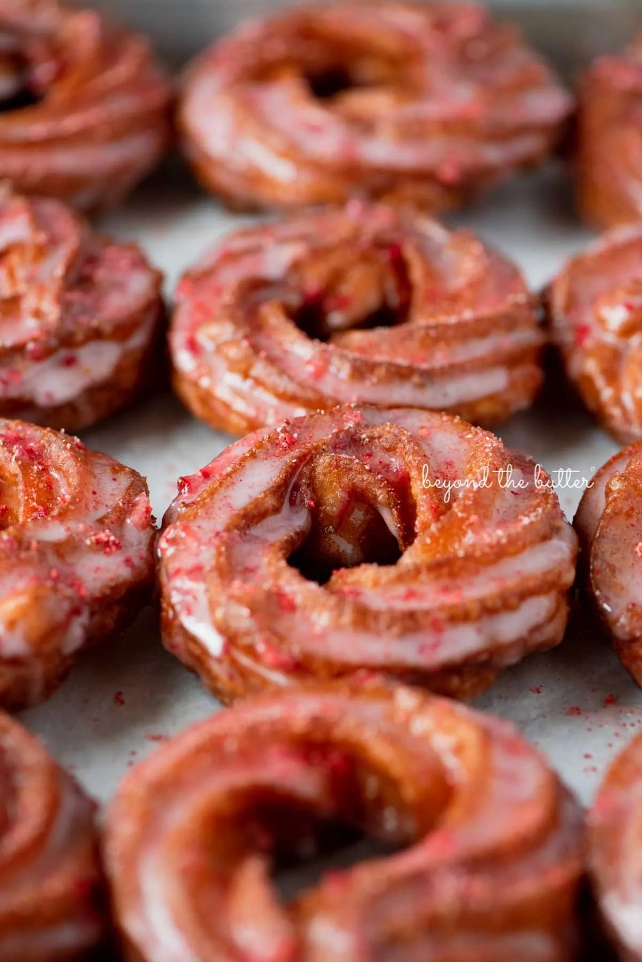 Strawberry glazed french crullers sprinkled with strawberry sugar on a parchment paper lined baking sheet | © Sovuh