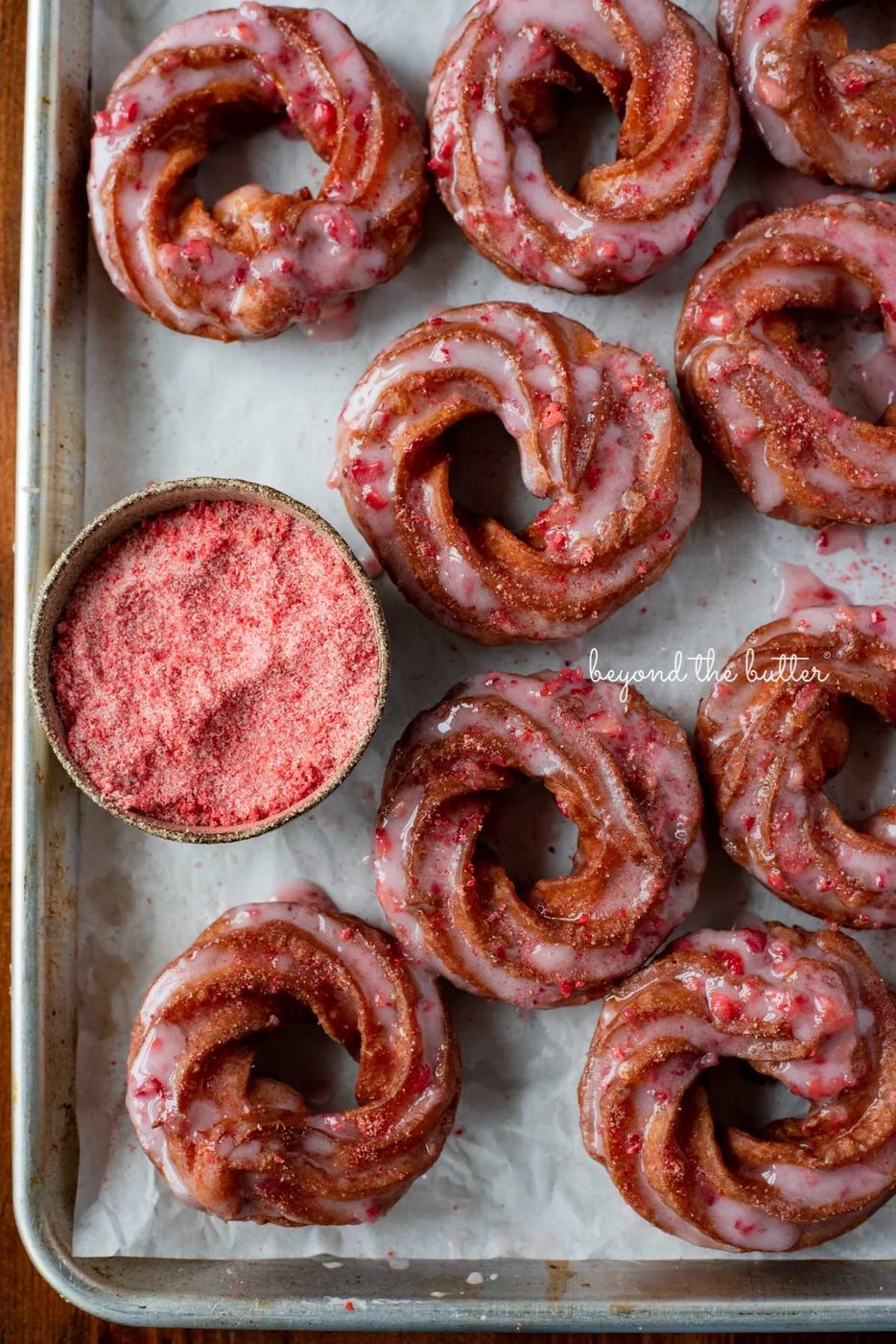 Strawberry glazed french crullers sprinkled with strawberry sugar on a parchment paper lined baking sheet | © Sovuh