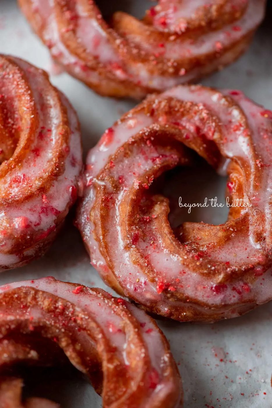 Strawberry glazed french crullers sprinkled with strawberry sugar on a parchment paper lined baking sheet | © Sovuh