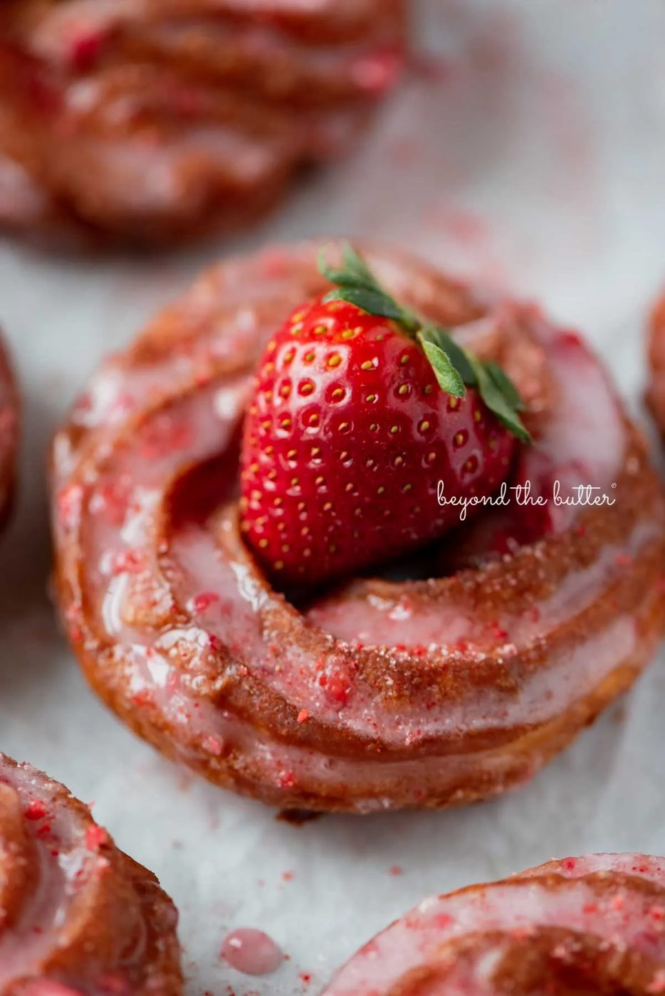 Just fried strawberry glazed french crullers on a parchment paper lined baking sheet topped with a fresh strawberry | © Sovuh