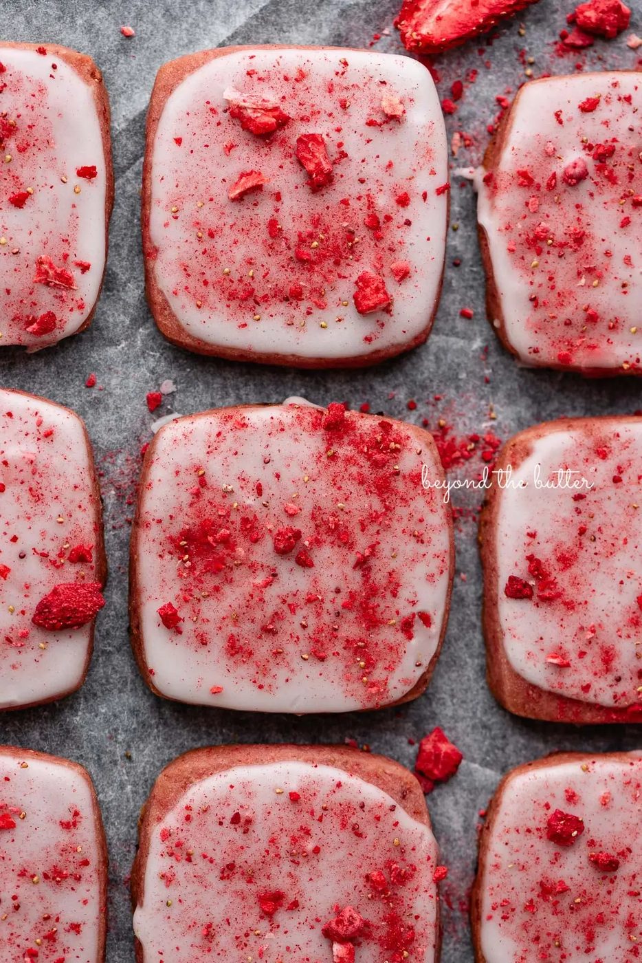Rows of strawberry shortbread cookies topped with a powdered sugar glaze and freeze-dried strawberry bits on a parchment paper and black background.
