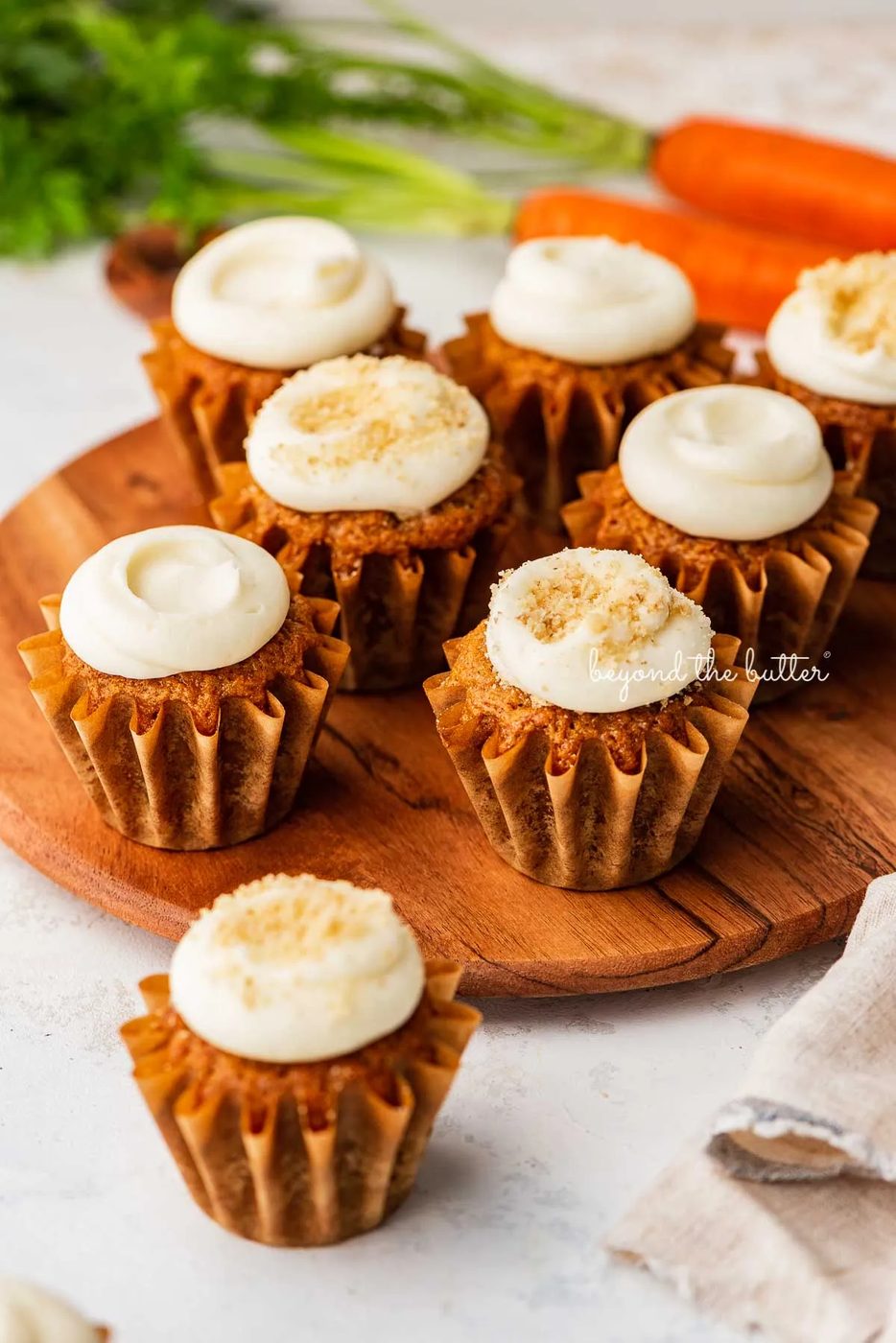 Super tender carrot cake cupcakes with cream cheese frosting placed on a wood cake stand on a white background with some carrots | © Sovuh