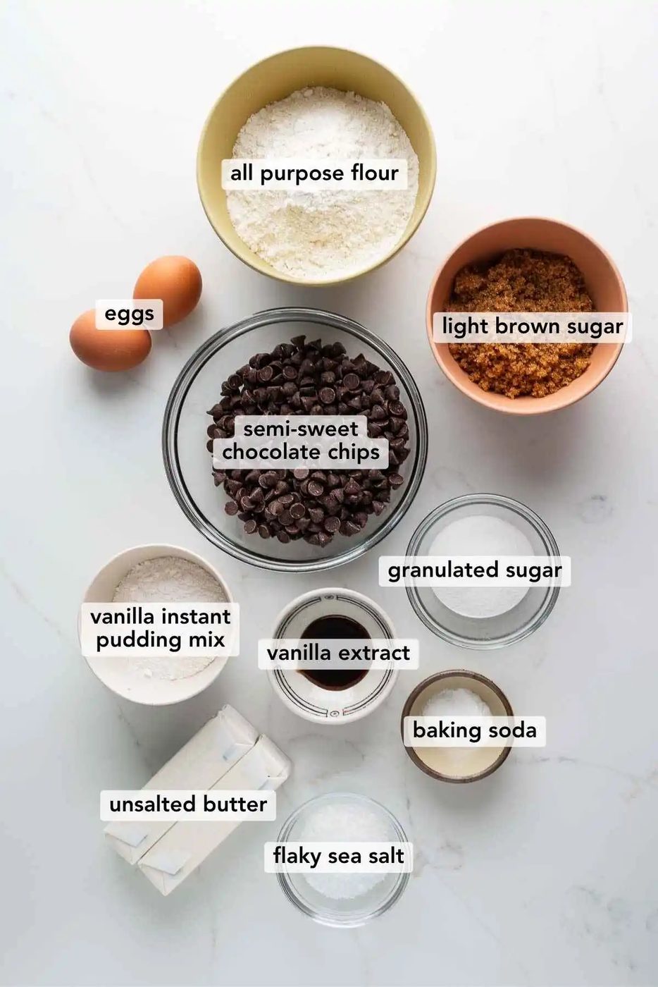Ingredients in various size bowls labeled as all purpose flour, eggs, light brown sugar, semi sweet chocolate chips, granulated sugar, vanilla instant pudding mix, vanilla extract, baking soda, unsalted butter, and flaky sea salt instant on a white marbled background.